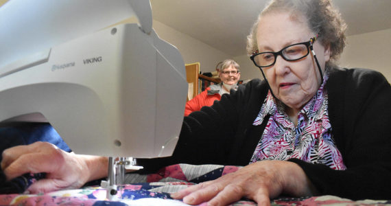 At her Enumclaw home on Feb. 22, Naomi Allen works on her latest quilt to be delivered to seniors in town. Her friend Janine Carpenter, in the background, talks to Allen regularly and helps deliver the quilts to the seniors who need them. Photo by Alex Bruell