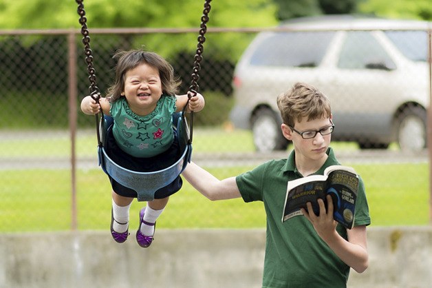 A boy multitasks while he pushes a small girl on the swings.
